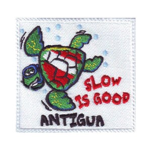 May include: A white embroidered patch with a cartoon turtle wearing a red, white, and green shell. The turtle is swimming and the text "Slow is Good Antigua" is embroidered in red and white.