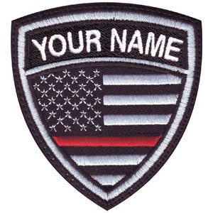 May include: Black and white embroidered patch with a red stripe. The patch features a shield shape with a US flag design and the text "YOUR NAME" at the top.