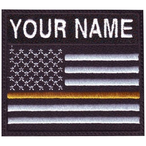 May include: Black and white embroidered American flag patch with a thin gold stripe across the middle. The text "YOUR NAME" is embroidered above the flag.
