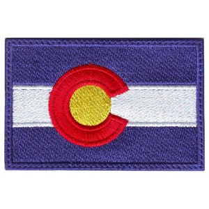 May include: An embroidered patch featuring the flag of Colorado. The flag is a blue field with a large, red circle containing a gold circle in the center. The circle is divided by a white line, creating a 'C' shape. The flag is surrounded by a white border.