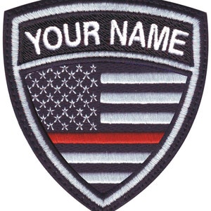 May include: Embroidered patch with a shield shape, featuring the text "YOUR NAME" above a stylized American flag design. The flag includes white stars, white and red stripes, and a red line.
