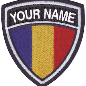May include: A black and white embroidered patch with a shield shape. The shield features the colors of the Romanian flag: blue, yellow, and red. The text "YOUR NAME" is embroidered in white on a black background above the shield.