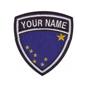 Peut inclure: Un écusson brodé bleu et blanc en forme de bouclier. L'écusson représente le drapeau de l'Alaska avec une grande étoile dorée et sept étoiles dorées plus petites. Le texte "YOUR NAME" est brodé en blanc sur fond noir en haut de l'écusson.