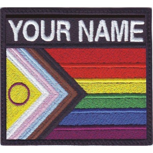 May include: A black and white embroidered patch with a rainbow flag design and the text "YOUR NAME". The flag features a yellow triangle with a purple circle in the center, followed by white, light blue, brown, pink, red, yellow, green, blue, and purple stripes.
