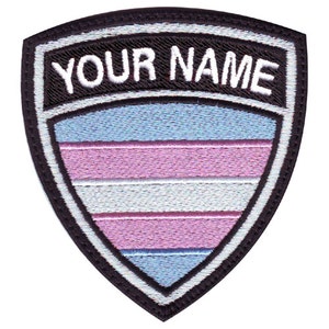 May include: A black and white embroidered patch in the shape of a shield with the text "YOUR NAME" at the top. The shield is filled with a transgender pride flag design with light blue, pink, and white stripes.