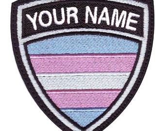 Può includere: Una toppa ricamata in bianco e nero a forma di scudo con il testo "YOUR NAME" in alto. Lo scudo è riempito con un design di bandiera del pride transgender con strisce blu chiaro, rosa e bianche.