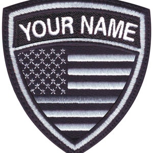 May include: Black and white embroidered patch with a shield shape. The shield has a white American flag design with the text "YOUR NAME" above it.