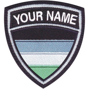 May include: A black and white embroidered patch with a shield shape. The shield has a green, white, and blue striped design. The text "YOUR NAME" is embroidered in white on a black banner above the shield.