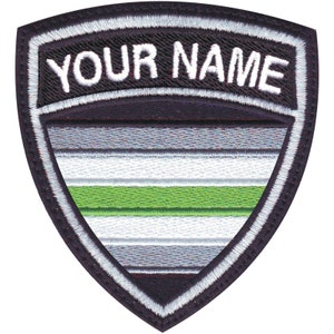 May include: A black and white embroidered patch in the shape of a shield with the text "YOUR NAME" at the top. The shield features a green, white, and gray striped design.
