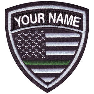 May include: Black and white embroidered patch with a thin green line, a shield shape, and the text "YOUR NAME" above an American flag.
