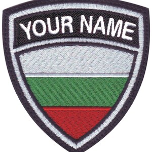 May include: Shield-shaped patch with the text "YOUR NAME" in white on a black background. Below, the patch features horizontal stripes in white, green, and red, bordered by a white stitched edge.