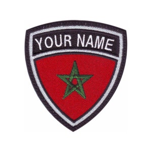 May include: A black and white embroidered patch with a red shield featuring a green five-pointed star. The text "YOUR NAME" is embroidered in white on a black banner above the shield.