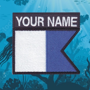 May include: A black and white embroidered patch with the text "YOUR NAME" in white. The patch features a blue and white flag with a white rectangle and a blue triangle.