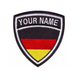 May include: Black and white embroidered patch with a shield shape. The shield has a German flag design with black, red, and yellow stripes. The text "YOUR NAME" is embroidered in white on the top of the shield.