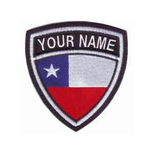 May include: A black and white embroidered patch with a shield shape. The shield features the Chilean flag with a white star on a blue background, a white stripe, and a red stripe. The text "YOUR NAME" is above the flag.