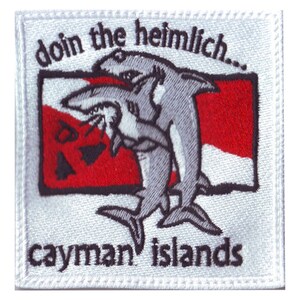 May include: White embroidered patch with a red and white diving flag. Two grey sharks are shown performing the Heimlich maneuver on each other. The text "doin the heimlich..." is above the sharks and "cayman islands" is below.