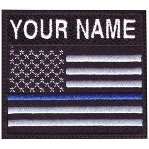 May include: Black embroidered patch with a white American flag and a blue thin line. The text "YOUR NAME" is above the flag.