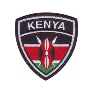May include: An embroidered patch with a black shield shape. The shield has a white border and the word "KENYA" in white letters. The center of the shield features the Kenyan flag, with red, black, and green stripes and a white spear design.