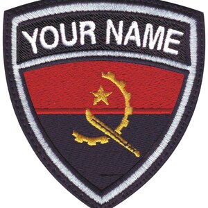 May include: A black and white embroidered patch with a red and black flag of Angola. The flag has a yellow star and a yellow symbol of a machete and a gear. The patch has a white border and the text "YOUR NAME" in white.
