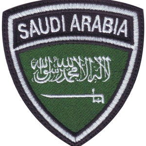 May include: A green and white embroidered patch with the words "SAUDI ARABIA" in white lettering on a black background. The patch features the flag of Saudi Arabia with the inscription of the Islamic declaration of faith in white.