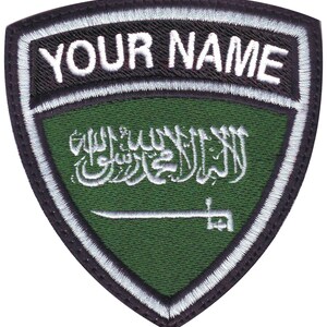May include: Embroidered patch with a shield shape. The top section features the text "YOUR NAME" in white. The main field is green with white Arabic script and a sword. The patch is bordered with black and white stitching.