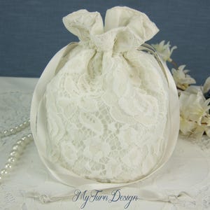Puede incluir: Una bolsa de encaje blanco con cordón, patrón floral y cierre de cinta de satén suave. La bolsa está decorada con un collar de perlas blancas y una rosa azul oscuro. El texto My Turn Design está impreso en azul en la parte inferior central.