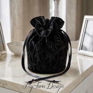 Könnte beinhalten: Ein schwarzer Samtbeutel mit Kordelzug und einem tonalen Damastmuster. Die Tasche hat einen gerafften Verschluss, der mit zwei langen schwarzen Satinbändern gesichert ist, die vorne herabhängen. Der Text My Turn Design ist unten schwarz aufgedruckt.