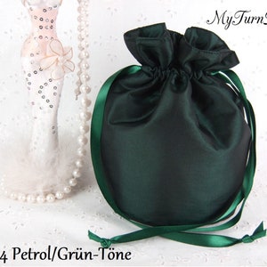 Può includere: Una borsa a cordoncino in raso verde scuro con un nastro verde. La borsa è mostrata su uno sfondo bianco con una collana di perle e una statuetta decorativa sullo sfondo. Il testo "4 Petrol/Grün-Töne" è visibile nell'immagine.