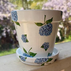 May include: White ceramic flower pot with a matching saucer, both decorated with blue hydrangea flower and green leaf designs. The pot is sitting on a white wooden surface, with a blurred background of trees and flowers.