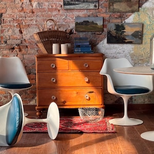 May include: A room scene featuring a wooden dresser with crystal knobs, a woven basket, and several small landscape paintings on a brick wall. White chairs with blue cushions and a small red rug complete the decor.