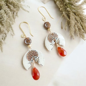 Könnte beinhalten: Goldfarbene Hängeohrringe mit floralem Design auf weißer, halbkreisförmiger Basis. Jeder Ohrring hat einen tropfenförmigen, orangeroten Edelstein und eine braune Perle. Die Ohrringe haben einen Hakenverschluss.