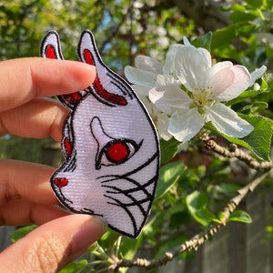 Peut inclure: Patch brodé blanc avec un motif de masque de renard rouge et noir. Le masque a des yeux rouges et un contour noir. Le patch est tenu dans une main sur fond d'arbre à pommes en fleurs.