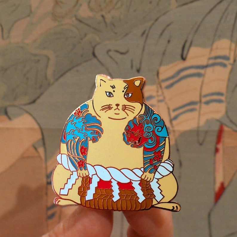May include: A gold-colored enamel pin of a cat wearing a blue and red patterned kimono, holding a brown and gold object in its paws. The cat has a white face with black eyes and a black nose.