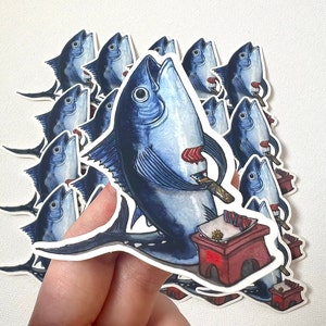 May include: A collection of stickers featuring a blue fish with a knife, a small red table, and sushi. The fish is depicted with its mouth open, holding a knife and sushi. The stickers have a white border.