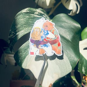May include: A sticker depicting a bear and a walrus wrestling. The bear is brown with tattoos, and the walrus is white with a yellow halo. They are wearing wrestling belts and surrounded by blue and red accents.