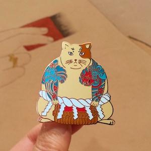 May include: A gold-colored enamel pin of a cat wearing a traditional Japanese sumo wrestler outfit. The cat is wearing a blue and red patterned loincloth and has a white and brown face.