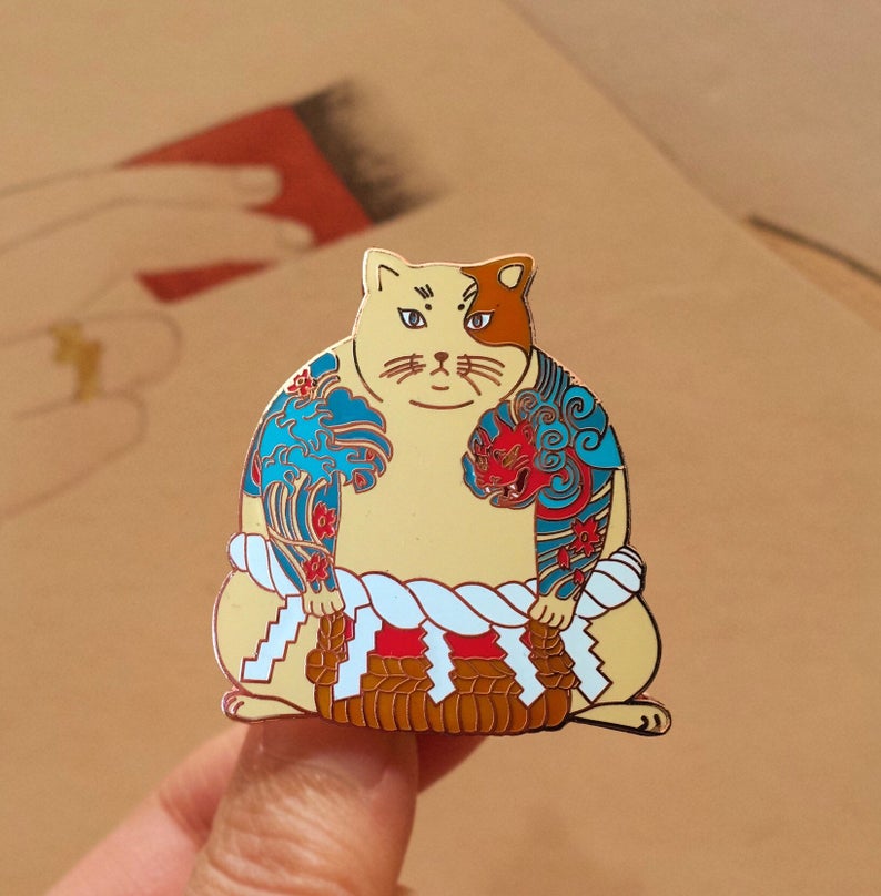 May include: A gold-colored enamel pin of a cat wearing a traditional Japanese sumo wrestler outfit. The cat is wearing a blue and red patterned loincloth and has a white and brown face.