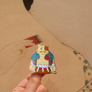 May include: A gold-colored enamel pin of a cat wearing a traditional Japanese sumo wrestler outfit. The cat has blue and red tattoos on its body and a white rope around its waist. The cat is sitting on a small, brown platform.