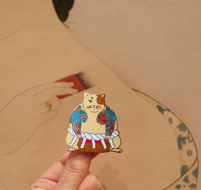 May include: A gold-colored enamel pin of a cat wearing a traditional Japanese sumo wrestler outfit. The cat has blue and red tattoos on its body and a white rope around its waist. The cat is sitting on a small, brown platform.