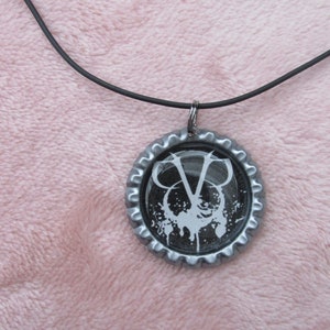 May include: A silver bottle cap necklace featuring a black and white graphic of a stylised 'V' with a splatter design.