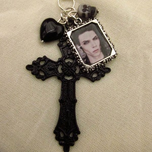 May include: A black gothic pendant with a cross, a framed portrait, a black heart, and a skull charm. The cross has an ornate design. The portrait is in a silver frame. The pendant is attached to a silver ring.
