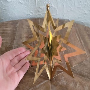 May include: A gold-coloured, multi-layered star ornament with a brass finish. The star has multiple points and a three-dimensional design. The ornament is likely for holiday or seasonal decor.