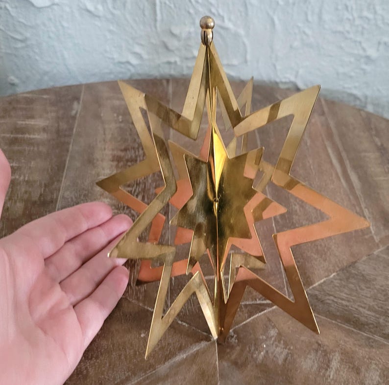 May include: A gold-coloured, multi-layered star ornament with a brass finish. The star has multiple points and a three-dimensional design. The ornament is likely for holiday or seasonal decor.