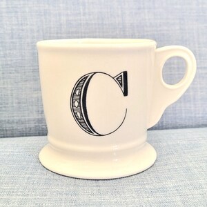 May include: White ceramic mug with a black outline of the letter "C" on the front.