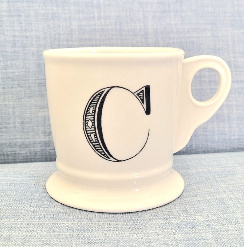 May include: White ceramic mug with a black outline of the letter "C" on the front.