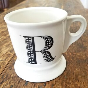May include: White ceramic mug with a black and white patterned letter R on the front. The mug has a handle and a wide base.