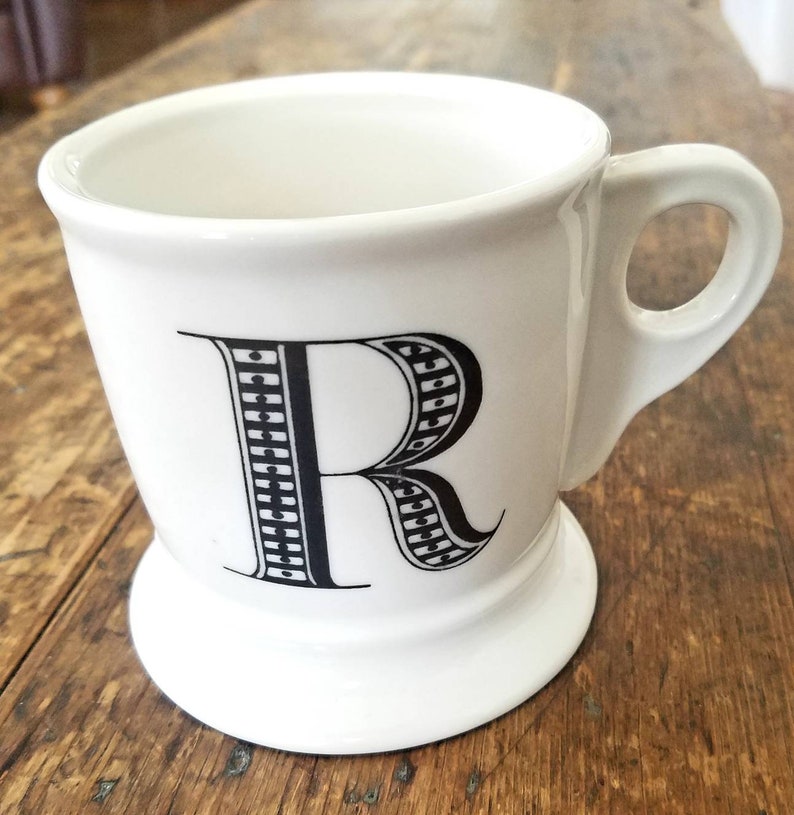 May include: White ceramic mug with a black and white patterned letter R on the front. The mug has a handle and a wide base.