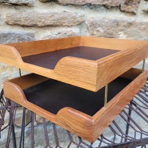 May include: A two-tiered desk organizer featuring two rectangular wooden trays with a natural light brown wood grain finish. Each tray has a dark brown interior base and is connected by thin, vertical brass-colored metal support rods.