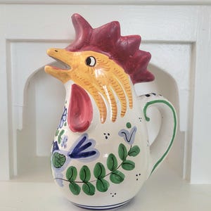May include: A ceramic pitcher in the shape of a rooster. The rooster has a yellow head, red comb and wattle, and a white body decorated with blue and green floral patterns. The handle is white with green trim.