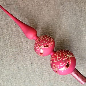 May include: A matte magenta glass Christmas tree topper featuring two stacked spherical ornaments decorated with gold glitter grid patterns and swirling lines, topped with a long, slender, pointed spire and a glittery base.